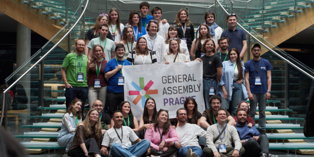 Participants at the General Assembly Prague pose together on a staircase, holding a large event banner inside a modern conference venue.